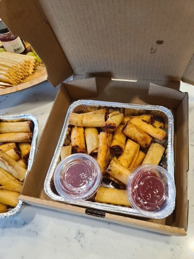 Open cardboard box containing lumpia with 2 small plastic containers of sauce.